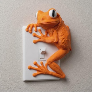 May include: A white rectangular light switch cover plate featuring a three-dimensional orange frog with textured skin, large white eyes with black pupils, and long toes gripping the edges of the plate while positioned next to the toggle switch.