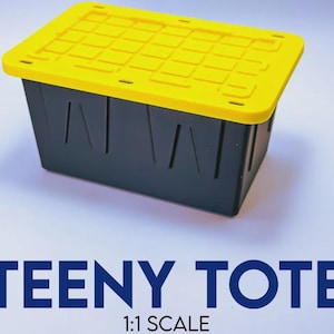 May include: A small rectangular storage bin with a solid black base and a bright yellow lid featuring a grid pattern. The text TEENY TOTE 1:1 SCALE is printed in large blue letters below the container, indicating its miniature size and scale.