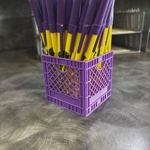 May include: A collection of yellow and purple pens stored upright in a small, square, purple plastic crate with a lattice design. The pens feature yellow barrels with purple caps and clips, and the crate has a distinct grid-like structure.
