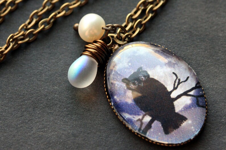 May include: A brass chain necklace with a glass pendant featuring a black silhouette of an owl perched on a branch against a starry night sky background. The pendant is accented with two white iridescent glass beads.