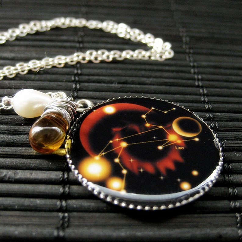 May include: A silver chain necklace with a round pendant featuring a black background with a constellation of stars and the word "Leo" in white. The pendant has a silver border with a beaded edge. The necklace also has a white pearl and a brown glass bead.