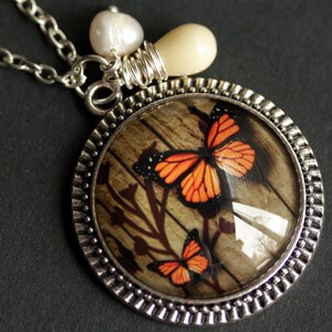 May include: A silver necklace with a round pendant featuring a wood-grain background with two orange and black butterflies. The pendant is surrounded by a silver beaded border. The necklace also includes a white pearl and a cream-colored bead.
