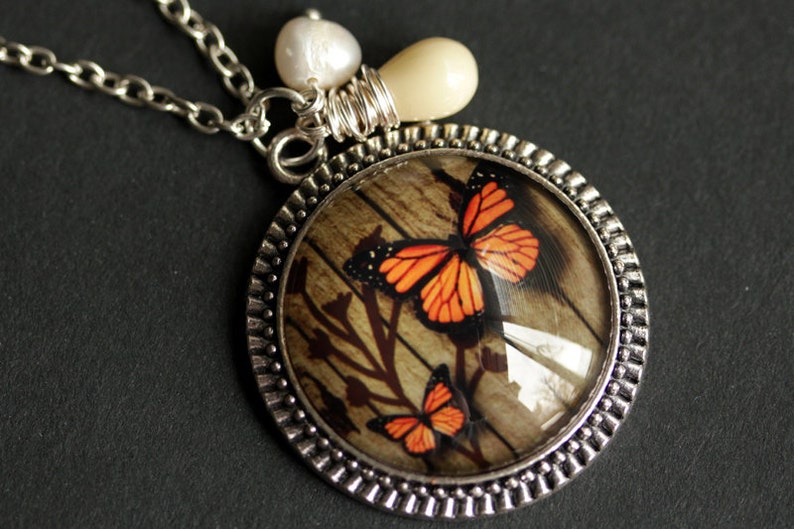 May include: A silver necklace with a round pendant featuring a wood-grain background with two orange and black butterflies. The pendant is surrounded by a silver beaded border. The necklace also includes a white pearl and a cream-colored bead.