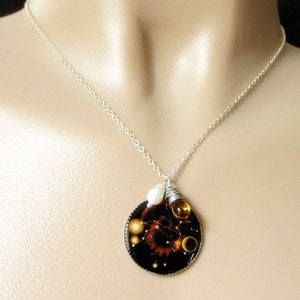 May include: A silver chain necklace with a round pendant featuring a black background with a constellation design and gold and white accents. The pendant is attached to a small pearl and a gold bead.
