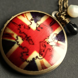 May include: A round locket necklace with a Union Jack design. The locket is made of metal and has a gold finish. The necklace is adorned with a white pearl and a black gemstone.