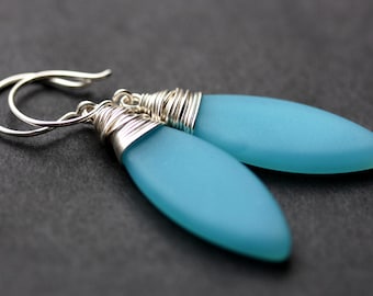 May include: A pair of aqua-colored, teardrop-shaped earrings. The earrings have a frosted glass appearance and are accented with silver wire wrapping and silver ear hooks. The earrings are displayed against a dark background.