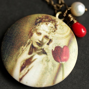 May include: A round locket necklace with a vintage image of a woman holding a red tulip. The locket is attached to a gold chain with a red and white bead and a white pearl.