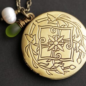 K&ouml;nnte beinhalten: Eine goldene Halskette mit einem Anh&auml;nger in Form eines Medaillons mit floralem Design. Das Medaillon ist mit einer Blume, Bl&auml;ttern und Spiralen verziert. Die Kette ist mit einer wei&szlig;en Perle und einer gr&uuml;nen Glasperle verziert.