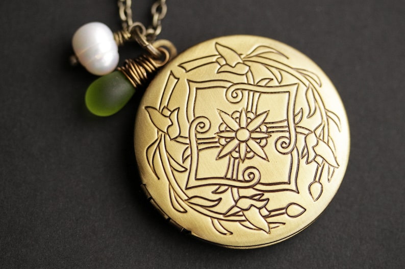 K&ouml;nnte beinhalten: Eine goldene Halskette mit einem Anh&auml;nger in Form eines Medaillons mit floralem Design. Das Medaillon ist mit einer Blume, Bl&auml;ttern und Spiralen verziert. Die Kette ist mit einer wei&szlig;en Perle und einer gr&uuml;nen Glasperle verziert.