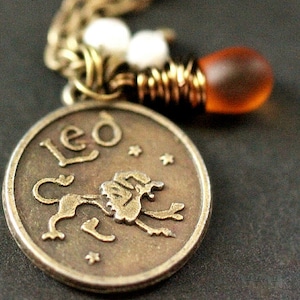 Op de afbeelding: Een bronzen Leo dierenriem hanger ketting. De ronde hanger toont het woord "Leo" en een leeuw illustratie. De ketting bevat een ketting, witte kralen en een bruine glazen kraal.