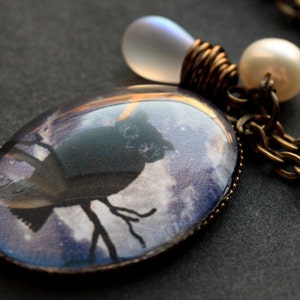 May include: A bronze chain necklace with a glass pendant featuring a silhouette of an owl against a purple and white cloudy sky. The pendant is attached to a clear glass bead and a white pearl.