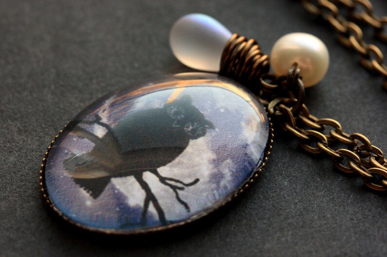 May include: A bronze chain necklace with a glass pendant featuring a silhouette of an owl against a purple and white cloudy sky. The pendant is attached to a clear glass bead and a white pearl.