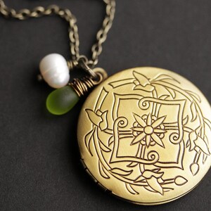K&ouml;nnte beinhalten: Eine goldene Halskette mit einem Anh&auml;nger in Form eines Medaillons mit floralem Muster. An dem Medaillon sind eine wei&szlig;e Perle und eine gr&uuml;ne Glasperle befestigt.