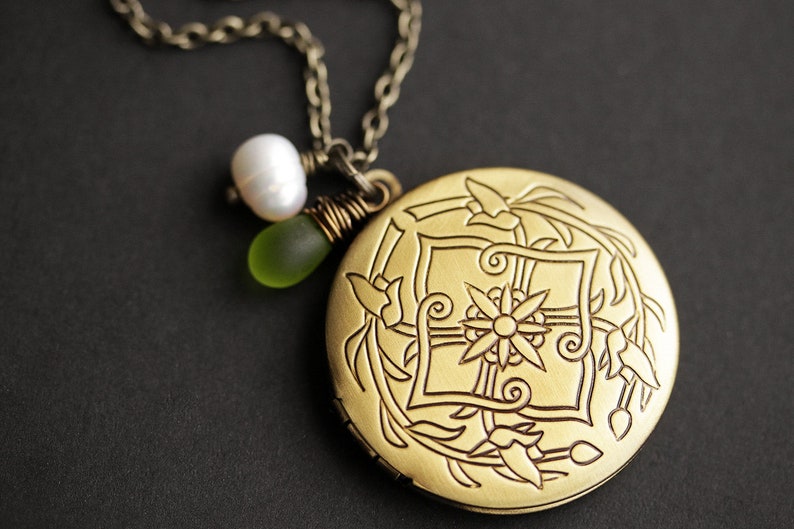 K&ouml;nnte beinhalten: Eine goldene Halskette mit einem Anh&auml;nger in Form eines Medaillons mit floralem Muster. An dem Medaillon sind eine wei&szlig;e Perle und eine gr&uuml;ne Glasperle befestigt.