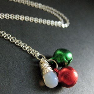 Puede incluir: Un collar de cadena de plata con un dije de campana blanca, verde y roja. Las campanas est&aacute;n unidas a un alambre de plata envuelto alrededor de una piedra blanca.