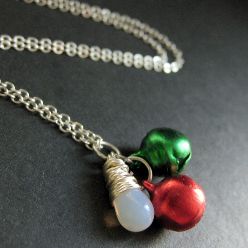 Puede incluir: Un collar de cadena de plata con un dije de campana blanca, verde y roja. Las campanas est&aacute;n unidas a un alambre de plata envuelto alrededor de una piedra blanca.