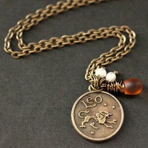 Op de afbeelding: Een bronskleurige ketting met een hanger met het woord "Leo" en een leeuwenillustratie. De ketting heeft een schakelontwerp en de ketting bevat witte kralen en een kleine amberkleurige glazen kraal.