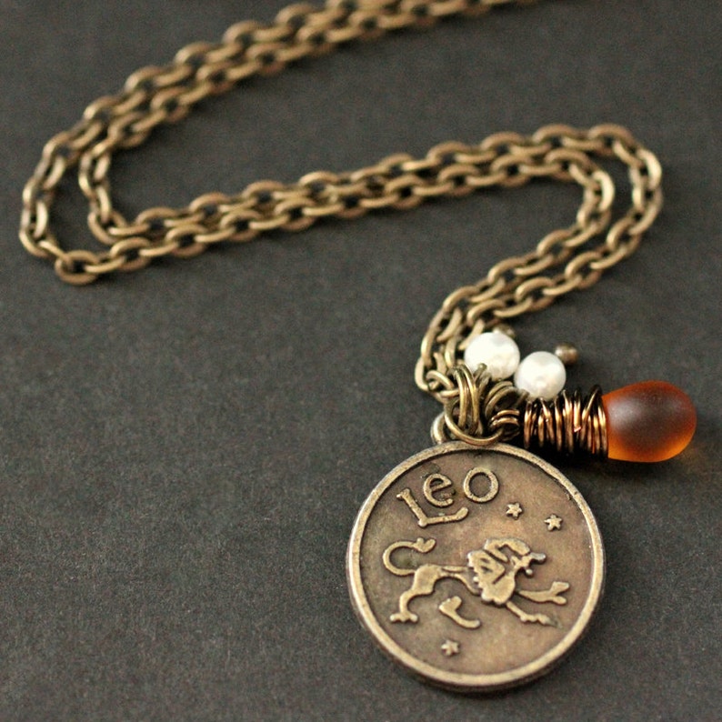 Op de afbeelding: Een bronskleurige ketting met een hanger met het woord "Leo" en een leeuwenillustratie. De ketting heeft een schakelontwerp en de ketting bevat witte kralen en een kleine amberkleurige glazen kraal.