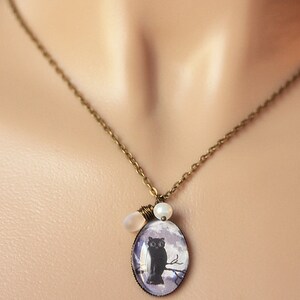 May include: A bronze chain necklace with a cameo featuring a black cat perched on a branch. The cameo is surrounded by two white pearl beads.