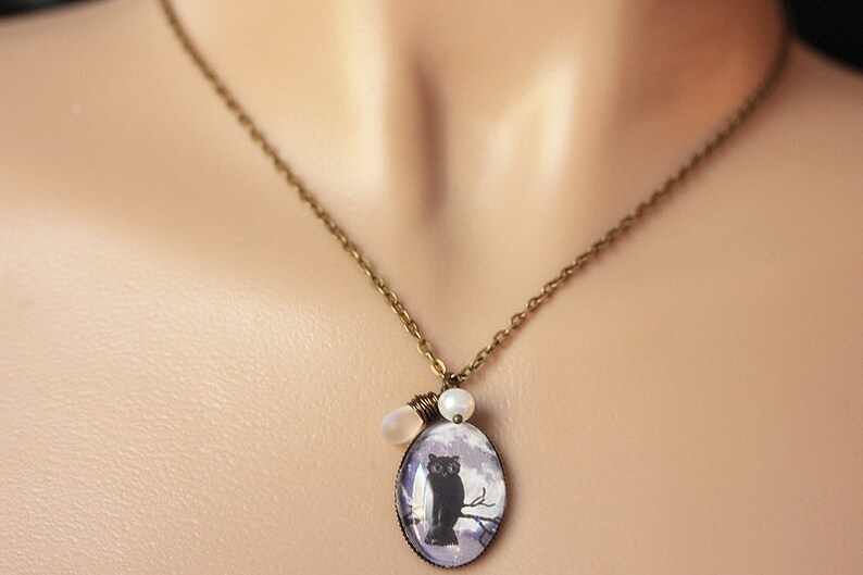 May include: A bronze chain necklace with a cameo featuring a black cat perched on a branch. The cameo is surrounded by two white pearl beads.