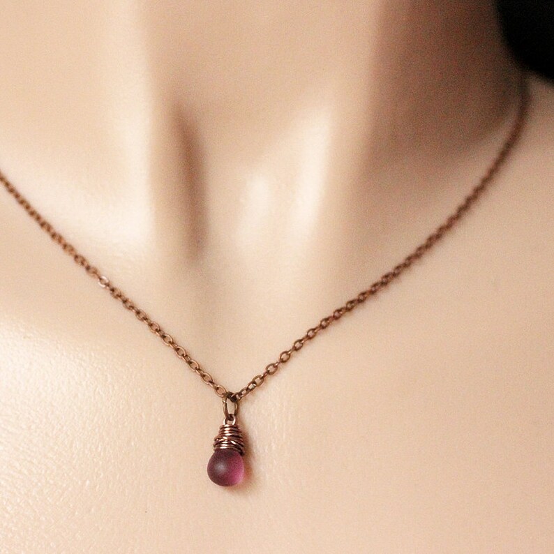 May include: A delicate copper chain necklace with a small, dark purple glass bead pendant. The bead is wrapped in copper wire.