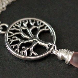Puede incluir: Un collar de plata con un colgante de árbol de la vida y una cuenta de vidrio rosa. El colgante es un círculo con un diseño de árbol detallado.
