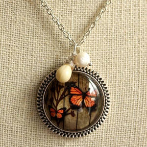 May include: A silver necklace with a round pendant featuring a vintage-style image of two orange butterflies on a woodgrain background. The pendant is accented with two white pearl beads.