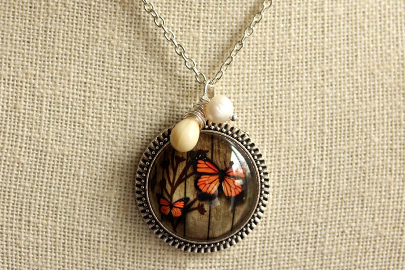 May include: A silver necklace with a round pendant featuring a vintage-style image of two orange butterflies on a woodgrain background. The pendant is accented with two white pearl beads.