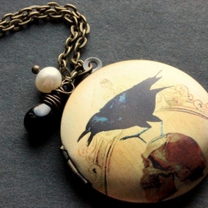 Puede incluir: Un collar de cadena dorada con un medallón redondo que presenta un cuervo negro posado sobre una calavera. El medallón tiene un encanto de perla blanca y una perla negra.