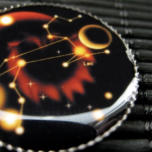 May include: A black and silver circular pendant with a red and orange image of the constellation Leo. The image also includes a crescent moon and stars.