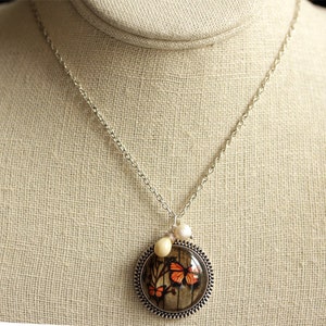 May include: A silver chain necklace with a round pendant featuring a butterfly design. The pendant has a brown and white background with orange butterflies. The necklace also has two small white beads.