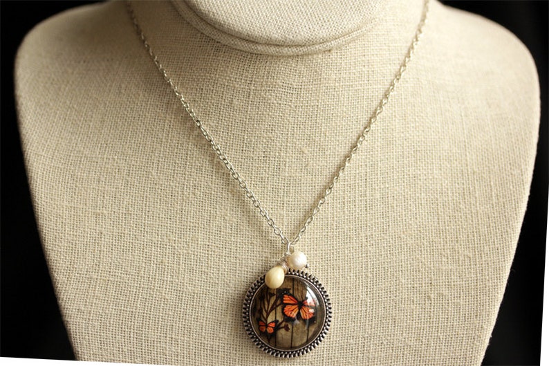 May include: A silver chain necklace with a round pendant featuring a butterfly design. The pendant has a brown and white background with orange butterflies. The necklace also has two small white beads.