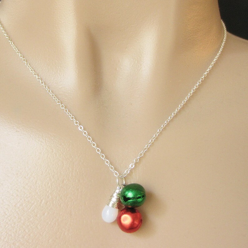 Puede incluir: Un collar de cadena de plata con un dije de tres cascabeles. Los cascabeles son rojos, verdes y blancos.