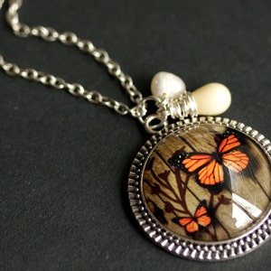 May include: A silver chain necklace with a round pendant featuring a woodgrain background and two orange butterflies. The pendant is accented with a white pearl and a silver charm.