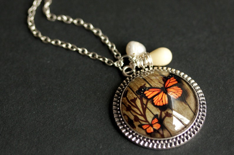 May include: A silver chain necklace with a round pendant featuring a woodgrain background and two orange butterflies. The pendant is accented with a white pearl and a silver charm.
