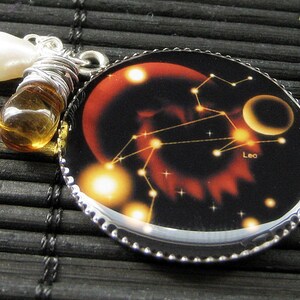 May include: A silver necklace with a round pendant featuring a black background with a red and orange nebula, a constellation of stars, and the word "Leo". The pendant is attached to a silver chain with a pearl and amber bead.