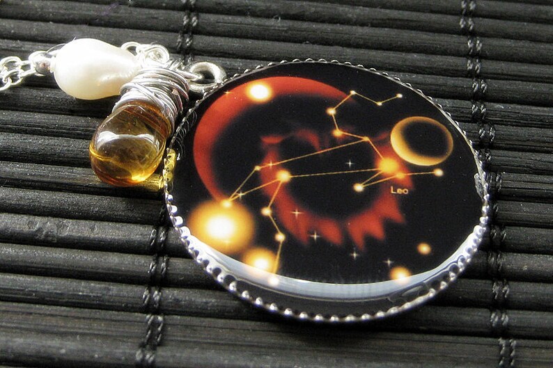 May include: A silver necklace with a round pendant featuring a black background with a red and orange nebula, a constellation of stars, and the word "Leo". The pendant is attached to a silver chain with a pearl and amber bead.