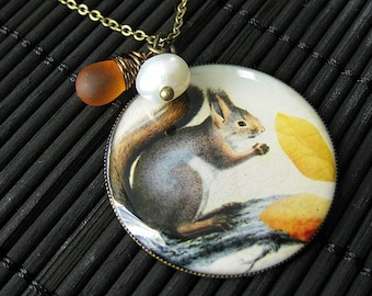 May include: A round pendant necklace with a squirrel and leaves design. The pendant is made of glass and has a gold-tone metal frame. The necklace is made of a gold-tone metal chain and has a brown glass bead and a white pearl bead.