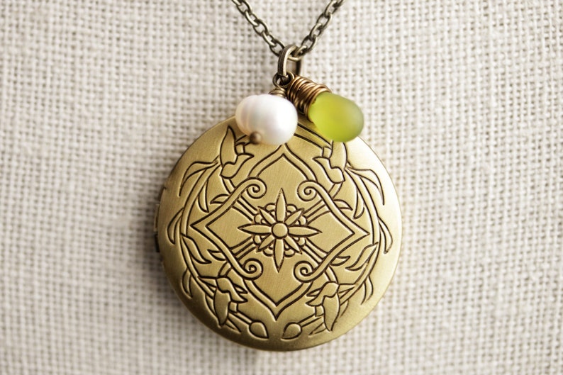 K&ouml;nnte beinhalten: Eine goldene Halskette mit einem detaillierten Blumenmuster. Das Medaillon hat eine wei&szlig;e Perle und eine gr&uuml;ne Glasperle daran.