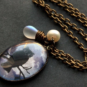 May include: A brass chain necklace with a pendant featuring a black silhouette of an owl perched on a branch, set against a purple and blue background. The pendant is surrounded by a clear glass dome. The necklace also features a white pearl and a clear iridescent glass bead.