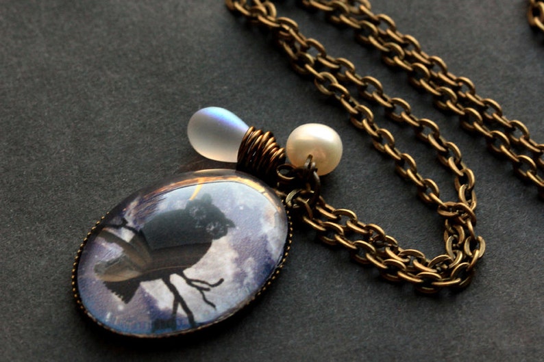 May include: A brass chain necklace with a pendant featuring a black silhouette of an owl perched on a branch, set against a purple and blue background. The pendant is surrounded by a clear glass dome. The necklace also features a white pearl and a clear iridescent glass bead.