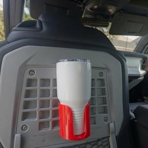 以下が含まれることがあります： 車両シートの背面にあるグレーのプラスチック製MOLLEグリッドパネルに取り付けられた、鮮やかな赤色のカップホルダーに白いタンブラーが収まっています。カップホルダーはグリッドに固定されており、飲み物を安定して収納できる構造になっています。