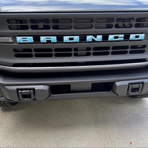 May include: The front grille of a vehicle features the word BRONCO in block letters. Each letter has a light blue background with a white wave pattern. The grille is matte black with horizontal slats and two black tow hooks on the bumper.