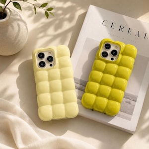 Puede incluir: Dos fundas para smartphone con una textura acolchada tridimensional brillante. Una funda es de color amarillo pastel pálido y la otra es de color verde lima vibrante. Ambas presentan un patrón de cuadrícula y un recorte para la cámara.