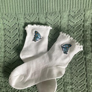 May include: A pair of white ribbed socks featuring a ruffled cuff and an embroidered blue butterfly with yellow accents on the upper section of each sock. The fabric has a vertical ribbed texture and the butterfly design is detailed in thread.