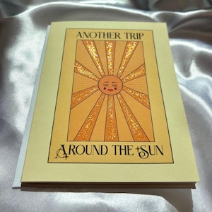 May include: A rectangular greeting card with a pale yellow background featuring a central illustration of a sun with a face, closed eyes, and glittery orange rays. The text reads ANOTHER TRIP AROUND THE SUN in black capital letters.