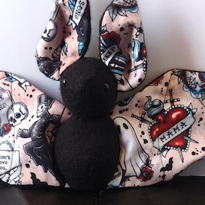 Könnte beinhalten: Ein Fledermaus-Stofftier mit einem Körper und Kopf aus schwarzem Filz. Die Flügel und Ohren bestehen aus hellrosa Stoff mit traditionellen Tattoo-Motiven, darunter rote Herzen mit dem Wort MAMA, Totenköpfe, Grabsteine, Geister und Schriftzüge.