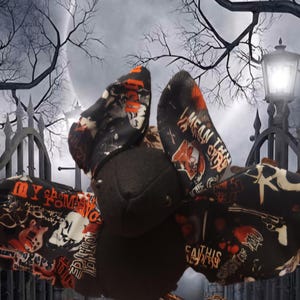 Könnte beinhalten: Ein Fledermaus-Plüschtier aus schwarzem Stoff mit einem sich wiederholenden Grafikdruck in Orange, Weiß und Rot. Das Muster enthält Texte wie 'MY CHEMICAL ROMANCE' und 'I MEAN THIS' sowie Abbildungen von Gitarren, Totenköpfen und Revolvern.