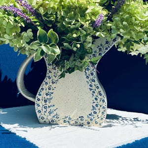 May include: A cream-colored ceramic pitcher with a speckled finish and a blue floral vine pattern along the edges. The pitcher is filled with a large arrangement of green hydrangea blossoms, purple flower spikes, and variegated ivy leaves.