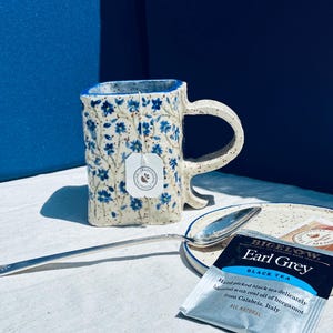 May include: A square-shaped ceramic mug with a speckled cream finish and blue floral pattern, featuring a handle and a tea bag inside. A silver spoon rests on a matching speckled saucer next to a Bigelow Earl Grey black tea bag packet.